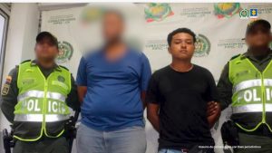 En la fotografía aparece un hombre de pie, moreno, cara redonda, cabellos negros crespos, de mediana estatura, con los brazos atrás esposado, vestido con una camiseta negra y un jean azul. Se encuentra custodiado por un uniformado de la Policía Nacional.