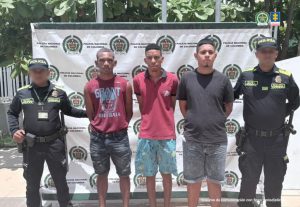 En la fotografía aparecen tres hombres jóvenes de pie, con los brazos atrás esposados, todos vestidos con camisetas y bermudas, custodiados por dos uniformados de la Policía Nacional.