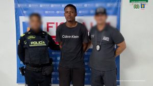 En la fotografía está un hombre de pie, de frente, alto, delgado, moreno con los brazos atrás esposados, cabellos negros, vestido con una camiseta, bermuda y chanclas negras. Se encuentra custodiado por un uniformado del Cuerpo Técnico de Investigación (CTI) y otro de la Policía Nacional. Detrás de ellos hay un pendón de la Fiscalía.