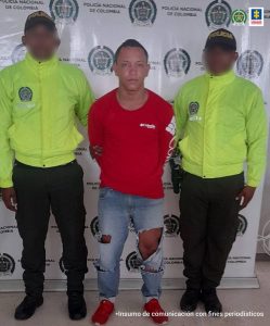 En la fotografía está un hombre de estatura baja, con los brazos atrás, moreno, contextura mediana, con los brazos atrás, vestido con una camiseta roja, un jean azul con rotos y tenis rojos. Se encuentra custodiado por dos uniformados de la Policía Nacional. Detrás de ellos hay pendón de esa institución. 
