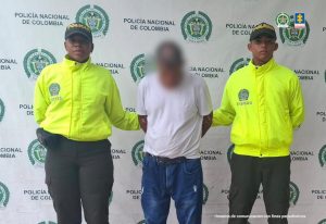 En la imagen está un hombre de pie, con la cabeza agachada difuminada, con los brazos atrás, vestido de camiseta blanca con jean azul y correa negra, custodiado por dos agentes de la Policía Nacional.