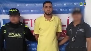 En la imagen está un hombre de pie, de frente, con los brazos atrás esposados, moreno, con cabellos cortos negros, barba negra, se encuentra custodiado por un uniformado del Cuerpo Técnico de Investigación (CTI) y un uniformado de la Policía Nacional. Detrás de ellos hay un pendón de la Fiscalía.