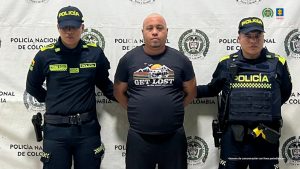  En la fotografía aparece un hombre blanco de pie, de frente, de contextura gruesa, con los brazos atrás esposado, semi calvo, vestido con camiseta y pantalón negro. Se encuentra custodiado por dos uniformados de la Policía Nacional. 