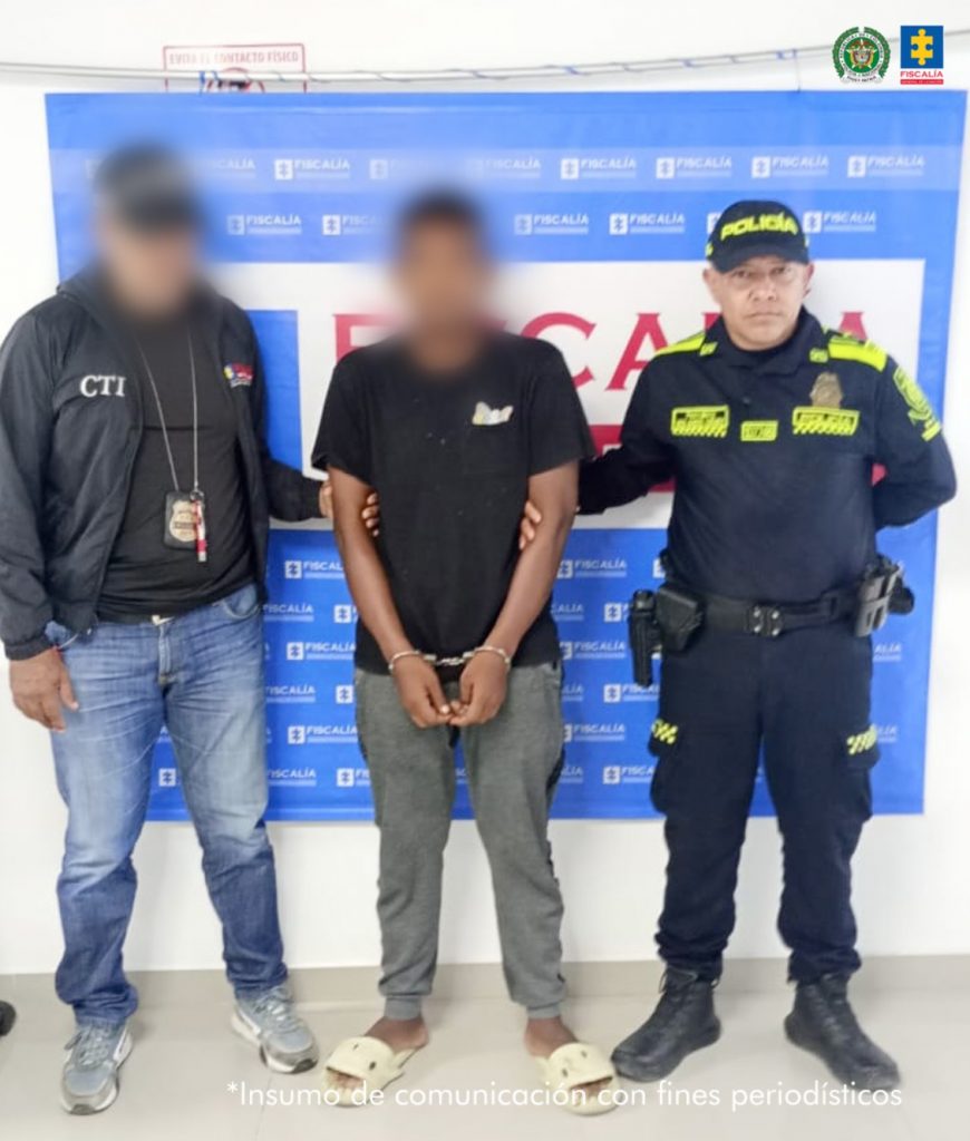 En la foto está un hombre de pie, con el rostro difuso, los brazos adelante esposados, vestido con camiseta negra, sudadera gris y sandalias blancas, custodiado por un uniformado del CTI y otro de la Policía Nacional.