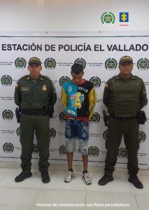 En la fotografía aparece un hombre de pie, con los brazos atrás esposado, vestido con un buzo de colores amarillo, negro y azul, bermuda de jean azul y tenis blanco.