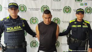 En la fotografía aparece un hombre de pie, con la cabeza agachada, cabellos negros, brazos atrás esposados, camiseta negra, custodiado por dos uniformados de la Policía Nacional. 
