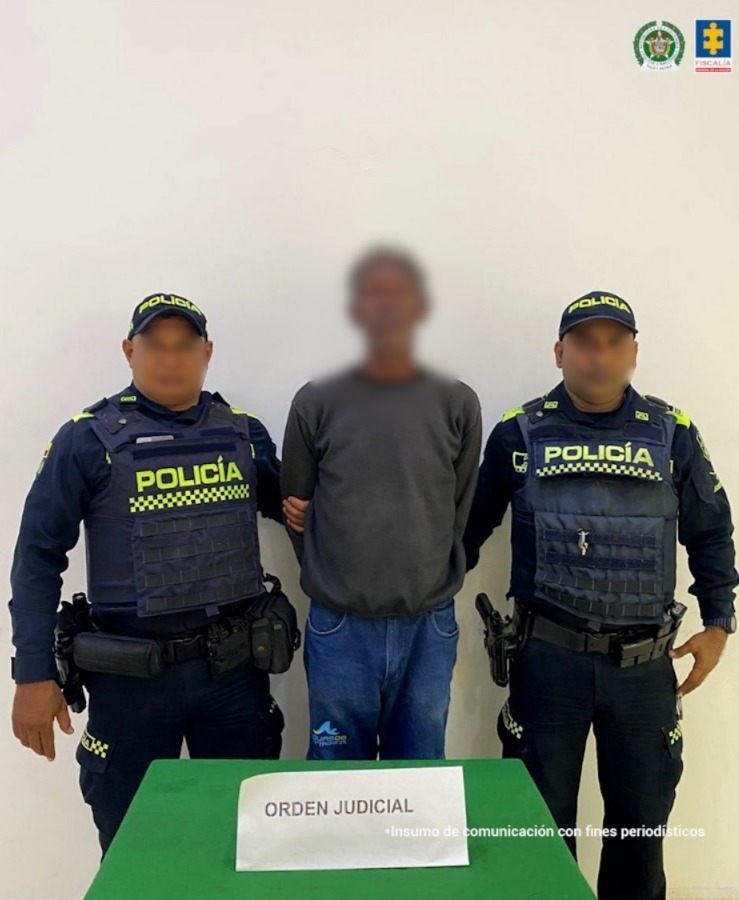 En la imagen está un hombre de pie, de frente, con los brazos atrás esposado, alto, moreno, delgado, vestido con un buzo manga larga gris y un jean azuly se encuentra custodiado por dos uniformados de la Policía Nacional. 