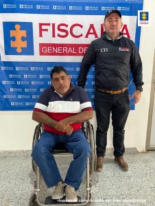 En la fotografía está un hombre sentado en una silla de ruedas, vestido con camiseta de rayas azul oscura, blanca y roja, jean azul y zapatos beige. Se encuentra custodiado por un uniformado del Cuerpo Técnico de Investigación, detrás de ellos hay un pendón de la Fiscalía.