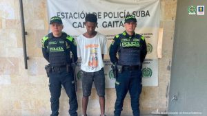 En la fotografía esta un hombre de pie, con la cabeza agachada, los brazos atrás, delgado, moreno, vestido de camiseta blanca y bermuda negra, custodiado por dos uniformados de la Policía Nacional.
