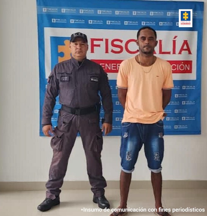 En la fotografía se observa a un hombre de tez morena, vestido de camiseta rosada con bermuda de jean, custodiado por un agente del CTI. Detrás de ellos se divisa un pendón de la Fiscalía General de la Nación.