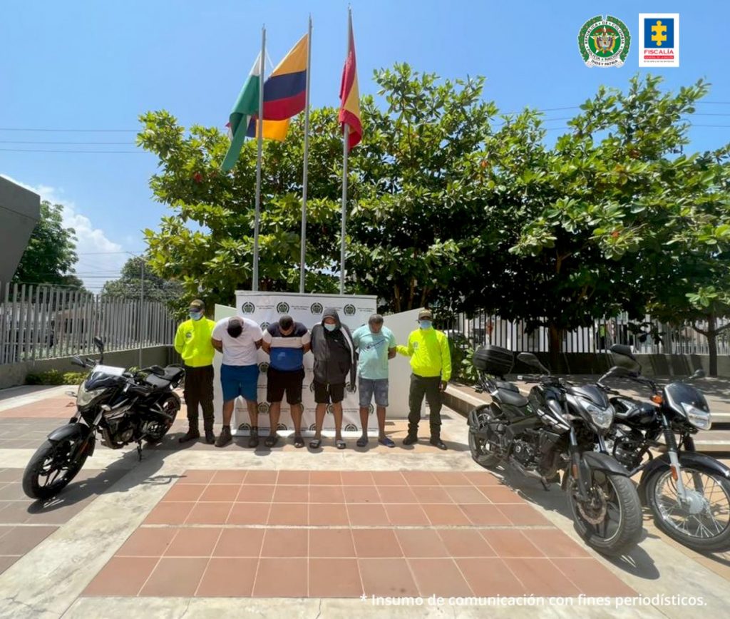 En la imagen se observa a cuatro hombres custodiados por dos agentes de la Policía Nacional, delante de un pendón de esa institución con las tres motos que les fueron incautadas.
