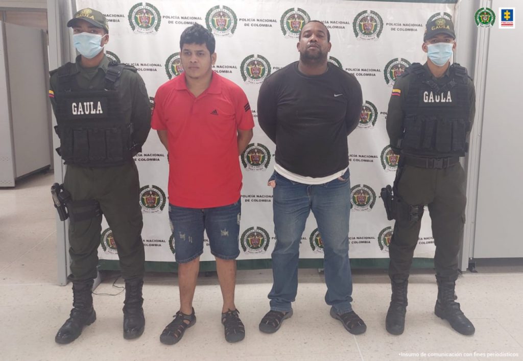 En la imagen se observa a dos hombres, uno de camiseta rojas y otro de buzo negro, custodiado por dos agentes de la Policía Nacional, delante de un pendón de esa institución