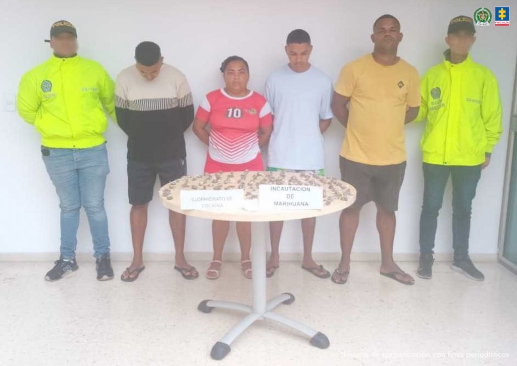 En la fotografía aparecen cuatro personas de pies, con los brazos atrás esposados, custodiados por dos uniformados de la Policía Nacional. Delante de ellos hay una mesa redonda con el estupefaciente incautado.