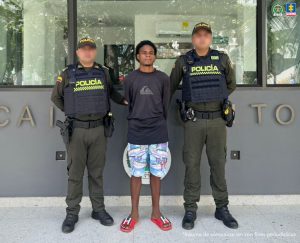 En la imagen, está un hombre moreno, de pie, con los brazos atrás, de cabellos negros crespos con medio afro, chivera, vestido con un buzo negro, bermuda azul estampada, sandalias rojas, custodiado por dos uniformados de la Policía Nacional.