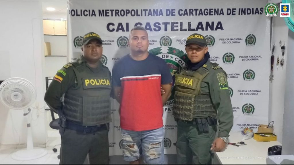 En la imagen se observa a un hombre de camiseta roja con azul oscuro y jean, custodiado por dos agentes de la Policía Nacional, delante de un pendón de esa institución.