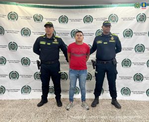 En la foto está un hombre trigueño, de pie, de frente, con los brazos atrás esposados, de mediana estatura, está vestido con camiseta roja, jean azul y tenis grises. Se encuentra custodiado por dos uniformados de la Policía Nacional. 
