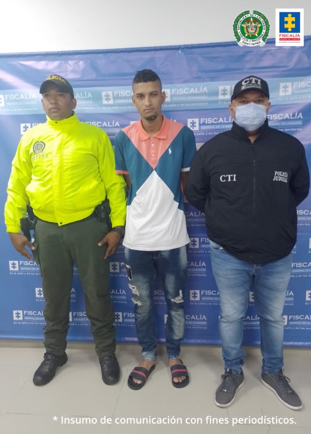 En la imagen se observa a un hombre delgado, moreno, vestido de camiseta blanca con colores azul y rosada, custodiado por un uniformado del CTI y por un uniformado de la Policía Nacional, delante de un pendón de la Fiscalía General de la Nación.