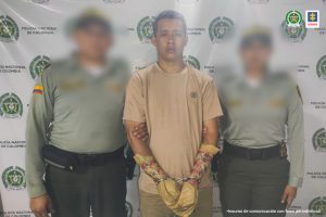 En la fotografía está un hombre de pie, color trigueño, con las manos esposadas, vestido de camiseta beige y pantalón verde, custodiado por dos agentes de la Policía Nacional. Detrás de ellos hay un pendón de esa institución.