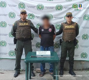 En la fotografía aparece un hombre de pie, de baja estatura con el rostro difuso, vestido de camiseta negra y jean, custodiado por dos agentes de la Policía Nacional. Delante de ellos hay una mesa con el arma blanca que se le encontró.