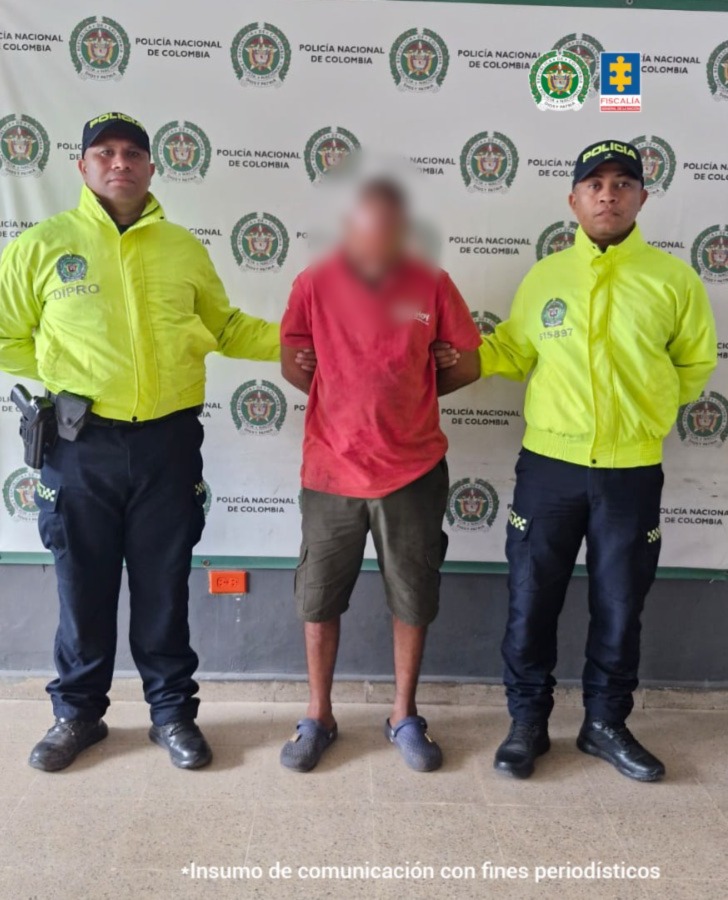 En la fotografía está un hombre moreno con el rostro difuso, de pie, con los brazos atrás esposados, custodiado por dos uniformados de la Policía Nacional. Se encuentra vestido con camiseta roja, bermuda verde y sandalias azules. 