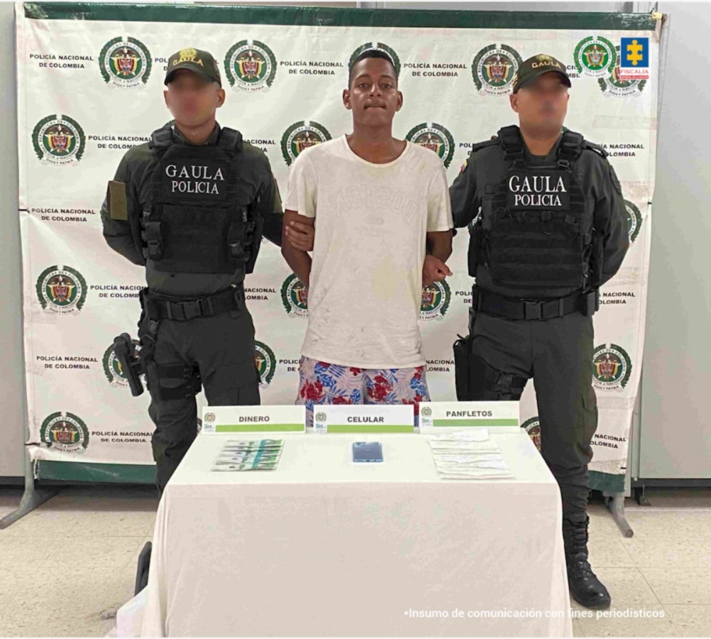 En la fotografía está un hombre joven de pie, moreno con los brazos atrás esposado, vestido con camiseta blanca y bermuda estampada de colores blanco, rojo y azul. Se encuentra custodiado por dos uniformados de la Policía Nacional.