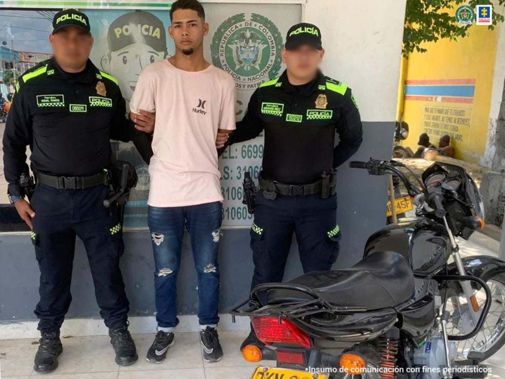 En la fotografía aparece un hombre vestido con camiseta rosada jean azul y tenis negro custodiado por dos agentes de la Policía Nacional. Detrás de ellos hay un pendón de esa institución y al lado se encuentra una motocicleta negra.