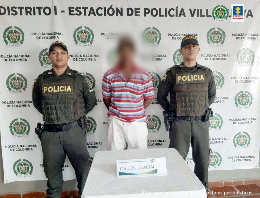 En la fotografía se observa a un hombre con el rostro difuso vestido de camiseta roja con azul y blanca y pantalón blanco custodiado por dos agentes de la Policía Nacional, delante de un pendón de esa institución.