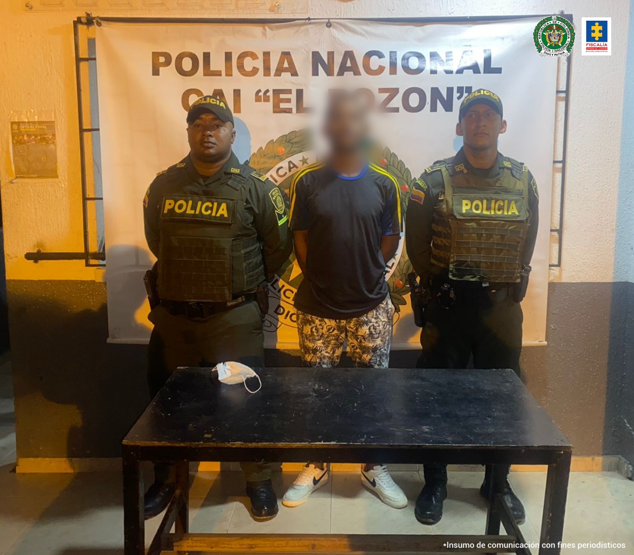 En la imagen se observa a un hombre de camiseta negra, bermuda y tennis custodiado por dos agentes de la Policía Nacional delante de un pendón de esa institución.