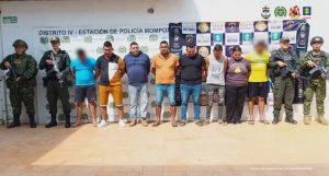  En la fotografía aparecen varios hombres de pie, parados con los brazos atrás, custodiados por uniformados de la Policía y la Armada Nacional. Detrás de ellos hay  pendones de esas instituciones y de la Fiscalía. 