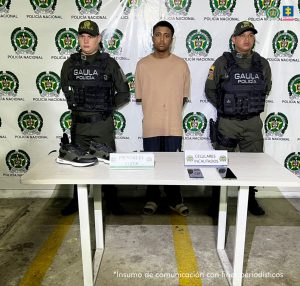  En la foto está un hombre joven, delgado, moreno, de pie, con los brazos atrás esposados, vestido con una camiseta beige, pantalón negro y sandalias blancas. Se encuentra custodiado por dos uniformados de la Policía Nacional. 