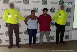 En la imagen están dos mujeres de pie, de contextura gruesa, mediana estatura, una se encuentra vestida de jean con camisilla blanca, sandalias café y la otra con camiseta roja, pantalón y sandalias gris. Se encuentran custodiadas por dos agentes de la Policía Nacional.