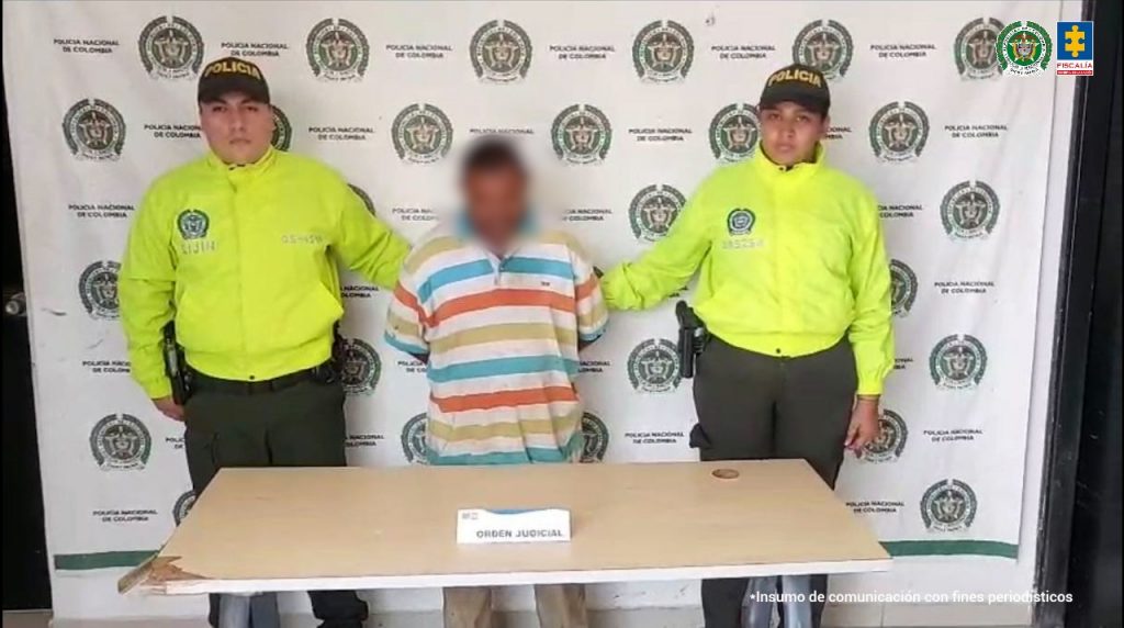 En la fotografía está un hombre de pie, de frente, con los brazos atrás esposados, con la cabeza agachada, el rostro difuminado, es de baja estatura y está vestido con una camiseta blanca con rayas azules, naranjas y beige. Se encuentra custodiado por dos uniformados de la Policía Nacional. 