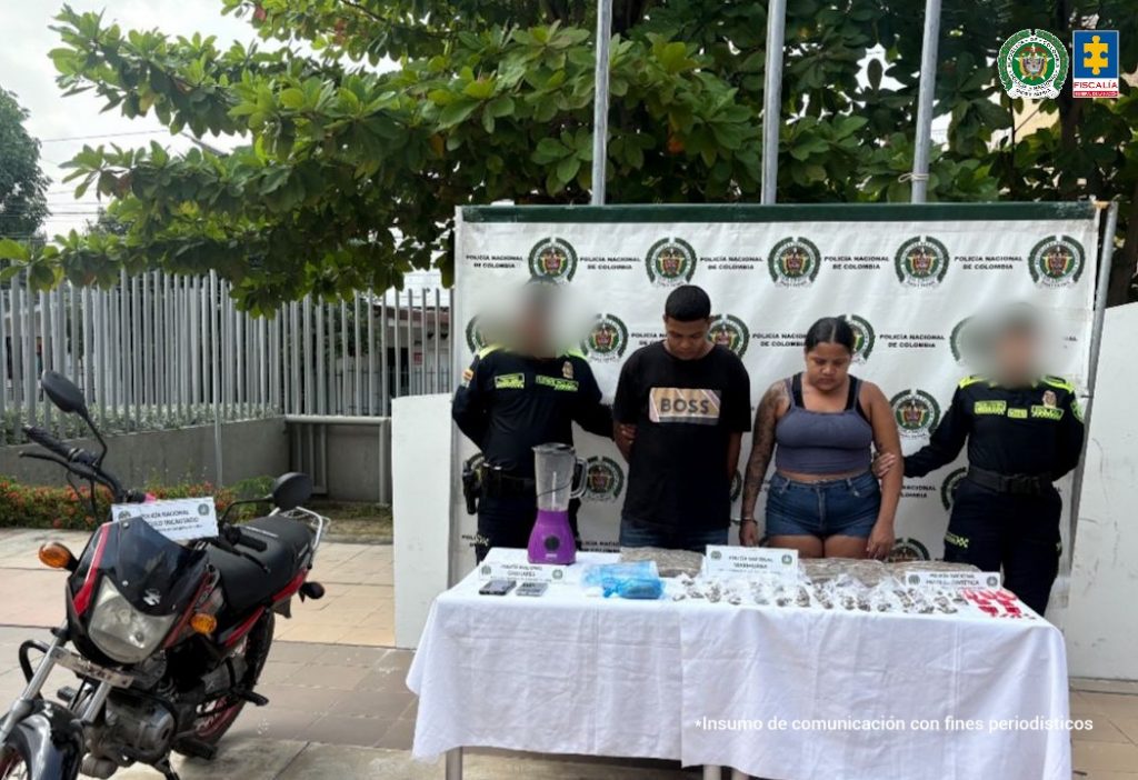 En la foto está un hombre y una mujer de pie, de frente, con las cabezas agachadas, ambas personas son morenas, la mujer de es de contextura mediana. El hombre está vestido con una camiseta negra y un jean azul y la mujer con una camisilla azul y una bermuda de jean azul. Se encuentran custodiados por uniformados de la Policía Nacional.