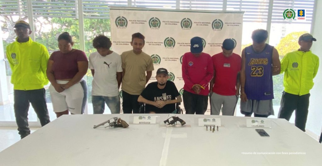 En la fotografía aparecen una mujer y seis hombres, uno está sentado en sillas de ruedas; se encuentran custodiados por dos agentes de la Policía Nacional. Delante de ellos hay una mesa donde están las armas y un celular incautado.