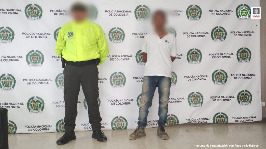 En la imagen aparece un hombre con camiseta blanca y jean custodiado por un agente de la Policía Nacional delante de un pendón de esa institución