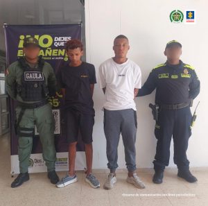 En la fotografía hay dos hombres jóvenes, de pie, con los brazos atrás, uno está vestido de camiseta y bermuda negra y el otro de camiseta blanca con sudadera gris. Se encuentran custodiados por dos uniformados de la Policía Nacional.