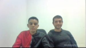 En la fotografía aparecen dos hombres morenos con cabellos cortos negros, se encuentran sentados al interior de una oficina asistiendo virtualmente a la audiencia en la que se le procesó.