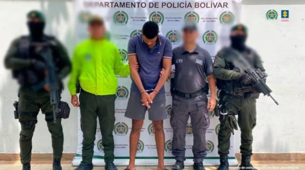 En la foto está un hombre delgado de pie, de frente, moreno, con la cabeza agachada, los brazos adelantes esposados, cabellos negros y se encuentra vestido con camiseta y bermuda azul. Uniformados del CTI y de la Policía aparecen custodiándolo.