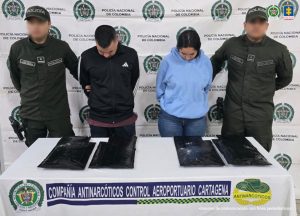 En la fotografía está un hombre y una mujer de pie, de frente, con las cabezas agachadas, los brazos atrás esposados, vestidos con sudaderas negras y azul respectivamente. A los lados se encuentran uniformados de la Policía Nacional y delante de ellos una mesa con la droga incautada.