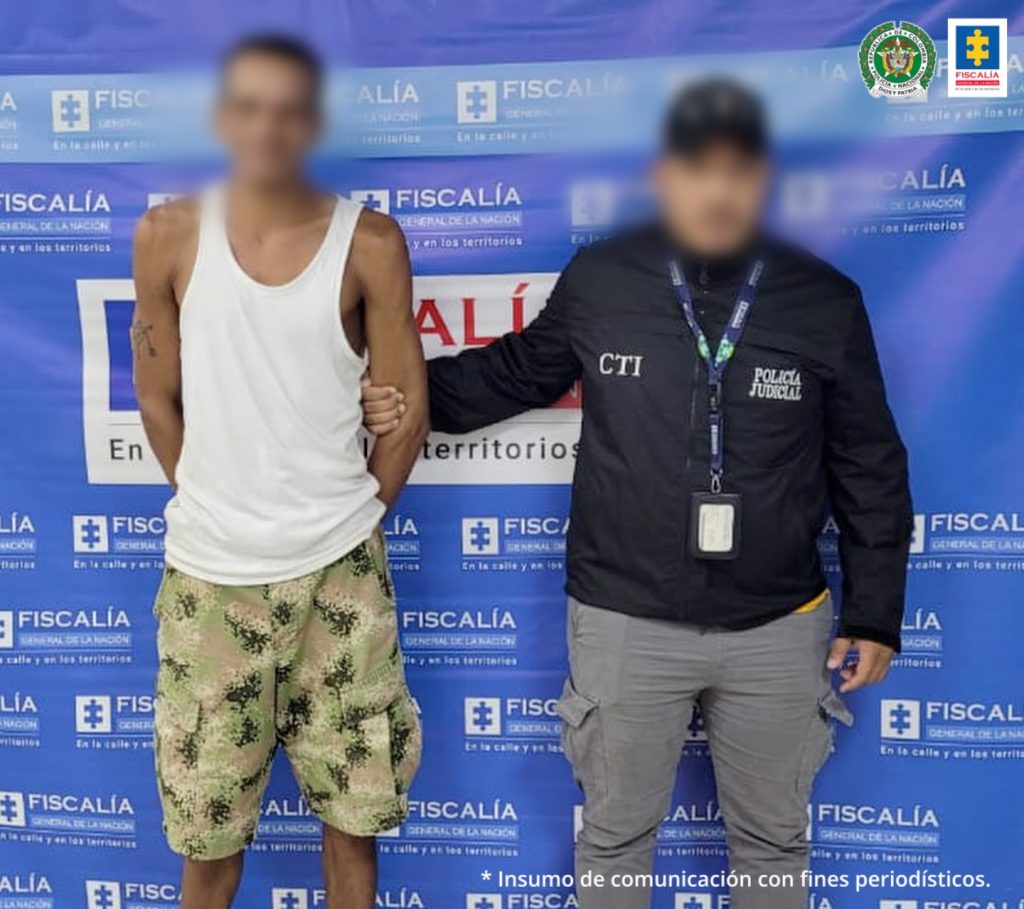 En la fotografía esta un hombre con el rostro difuso, se encuentra de pie, con los brazos atrás, es delgado y viste una camisilla blanca con una bermuda verde estampada. A su lado está un uniformado del CTI con el rostro difuso y detrás de ellos hay un pendón de la Fiscalía.