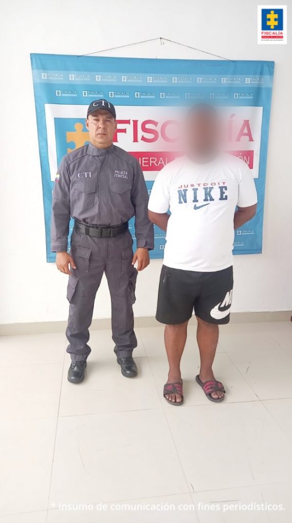 En la fotografía aparece un hombre moreno, vestido de camiseta blanca, bermuda negra y sandalias negras, custodiado por un servidor de CTI. Detrás de ellos hay un pendón de la Fiscalía General de la Nación.