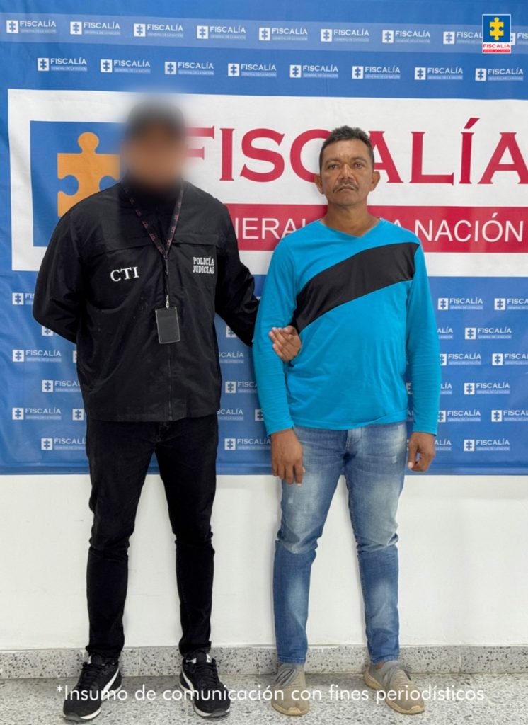 En la foto está un hombre moreno de pie, con bigote, cabellos negros ondulados, vestido con un buzo azul con negro, jean azul y tenis grises. Se encuentra custodiado por un uniformado del CTI. Detrás de ellos hay un pendón de la Fiscalía.