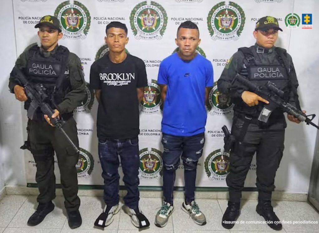 En la fotografía están dos hombres de pie, de frente, con los brazos atrás esposados, uno está vestido con camiseta, pantalón y tenis negros y el otro con camiseta azul, pantalón negro y tenis blanco. Se encuentran custodiados por dos uniformados de la Policía Nacional.