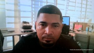  En la fotografía está el rostro de un hombre moreno, cabellos cortos negros crespos, tiene una pequeña barba en el mentón. Está sentado al interior de una oficina asistiendo virtualmente a la audiencia en la que se le procesó.