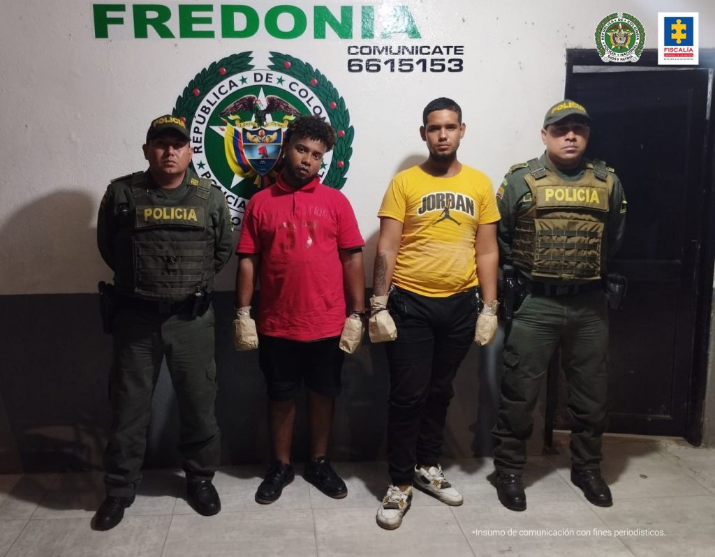 En la fotografía aparecen dos hombres morenos, uno vestido de camiseta roja, bermuda y tenis negros, el otro con camiseta amarilla, pantalón negro y tenis beige, custodiados por dos agentes de la Policía Nacional. Detrás de ellos hay una pared con el escudo de esa institución que dice CAI Fredonia.