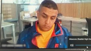 En la fotografía aparece el rostro de un hombre trigueño de cabellos cortos, boca delgada, nariz recta, vestido de camiseta amarilla con saco azul. Se encuentra sentado en una silla en el interior de una oficina asistiendo virtualmente a la audiencia en la que se le procesó.