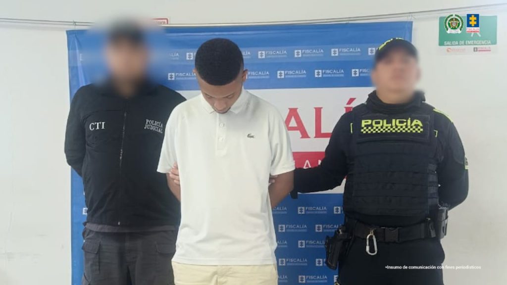 En la fotografía aparece un hombre moreno, de pie, de frente, con los brazos atrás esposados, con la cabeza agachada, de cabello corto, crespo negro. Está vestido con una camiseta blanca y custodiado por un uniformado del CTI y otro de la Policía Nacional.
