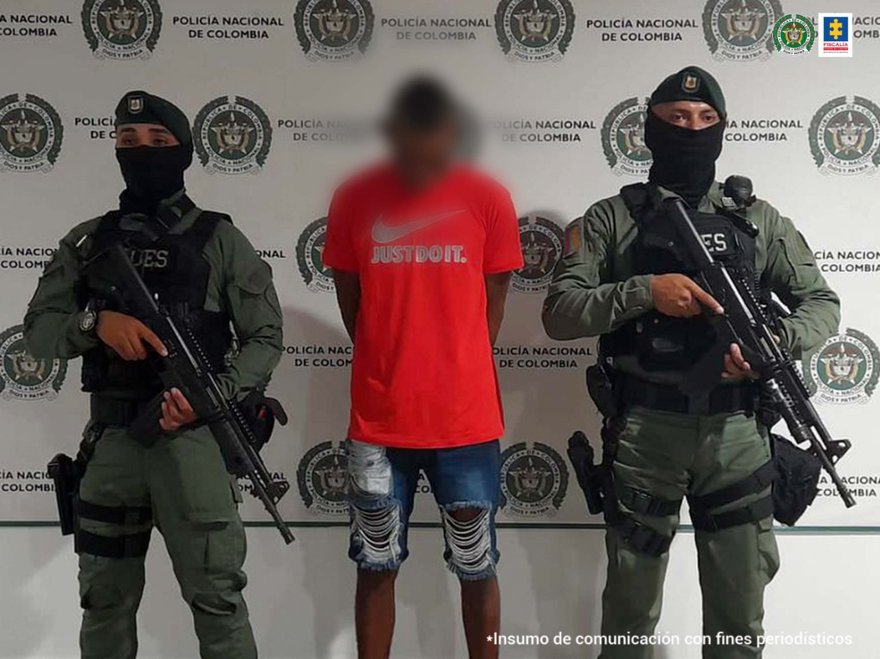 En la foto se observa a un hombre con camiseta roja y bermuda azul custodiado por dos agentes de la Policía Nacional con el fondo de un pendón de esa institución.