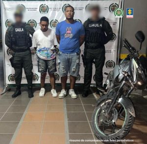 En la fotografía aparecen dos hombres de pie, con los brazos atrás, morenos, de contextura gruesa, uno de estatura media y el otro de estatura baja, custodiado por dos uniformados de la Policía Nacional.