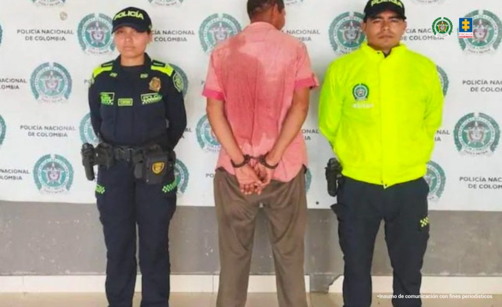 En la fotografía aparece un hombre alto, de pie, de espalda, con las manos esposadas, vestido con una camisa roja manga corta y un pantalón café. Se encuentra custodiado por dos uniformados de la Policía Nacional con un pendón de esa institución.